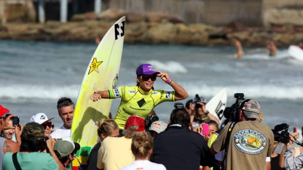 An image of Layne Beachley surrounded by cameras with a surfboard behind her