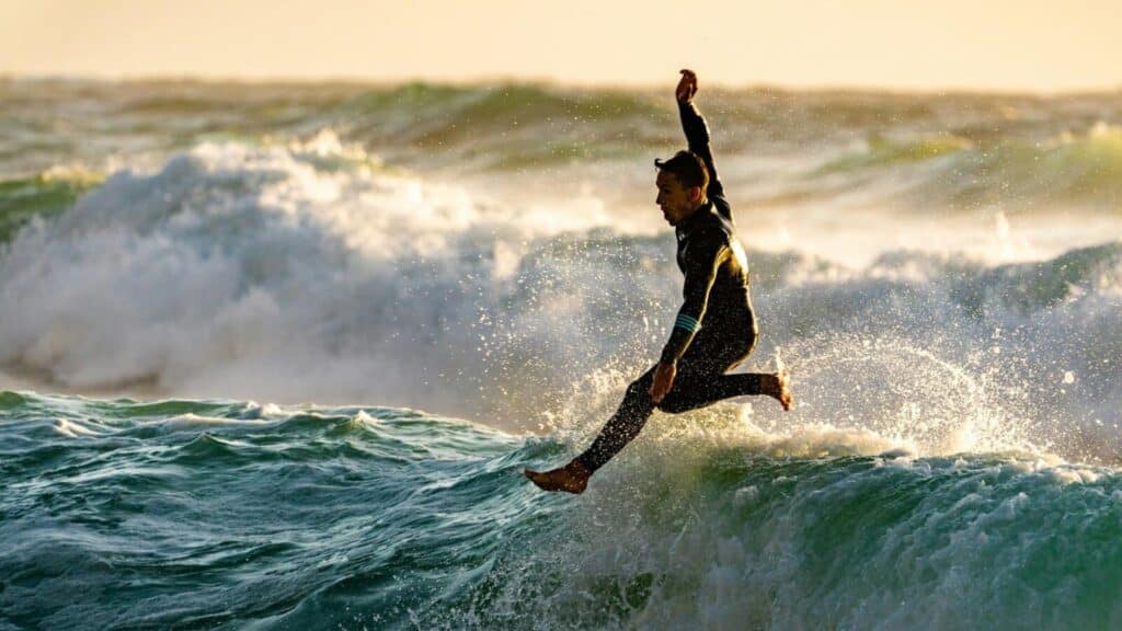 Ein Bild von einem Mann, der vom Surfbrett gefallen ist und gerade im Wasser landet. Er trägt einen Neoprenanzug  
